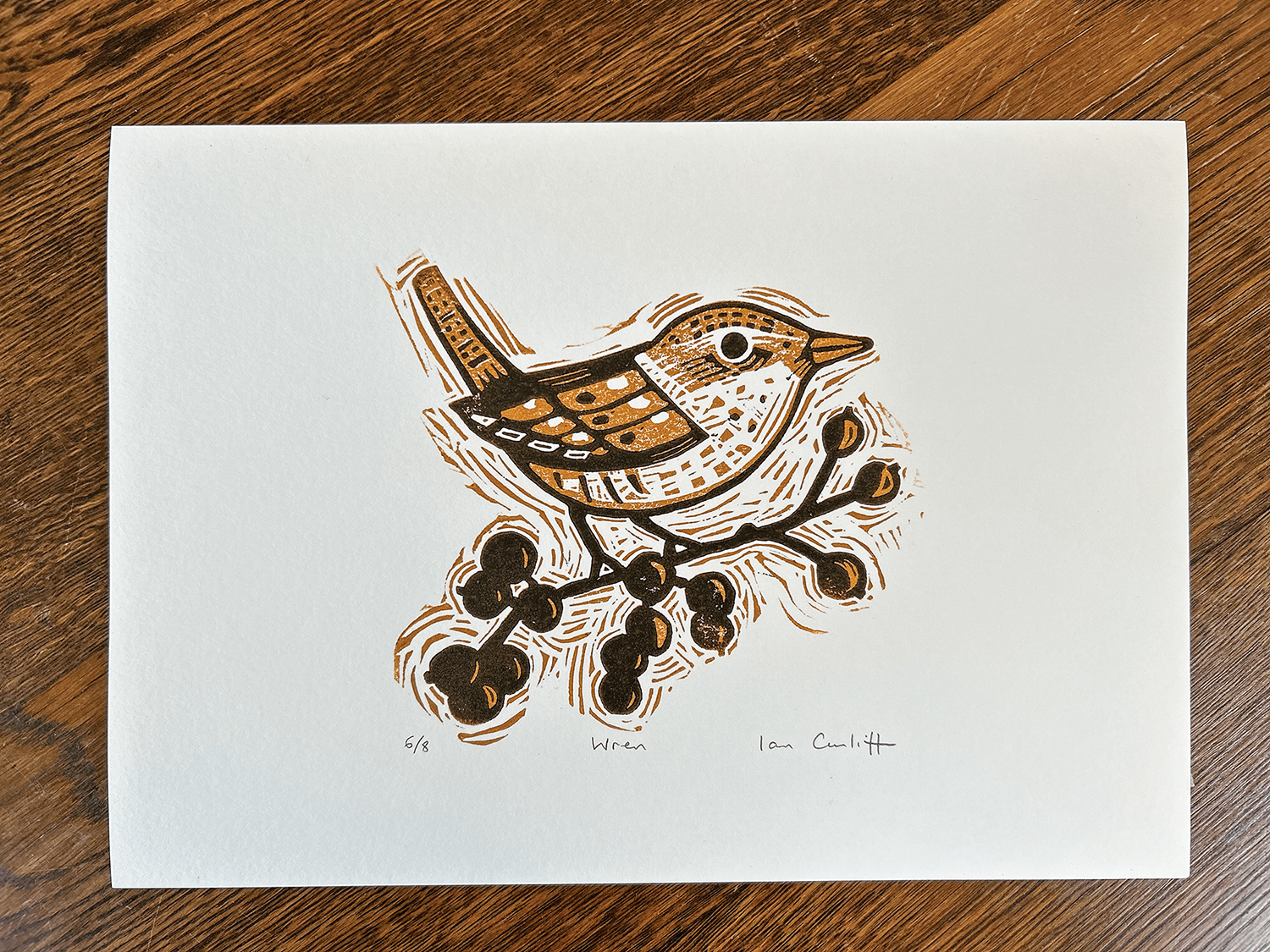 original linocut illustration of a bird on a branch with berries on a white sheet, placed on a wooden surface.