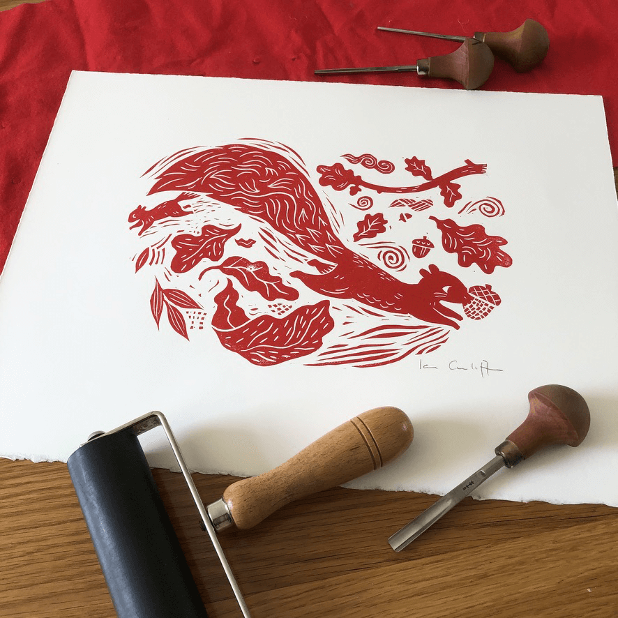 Red woodcut print of a squirrel and branches on white paper with printing tools on a red fabric background.